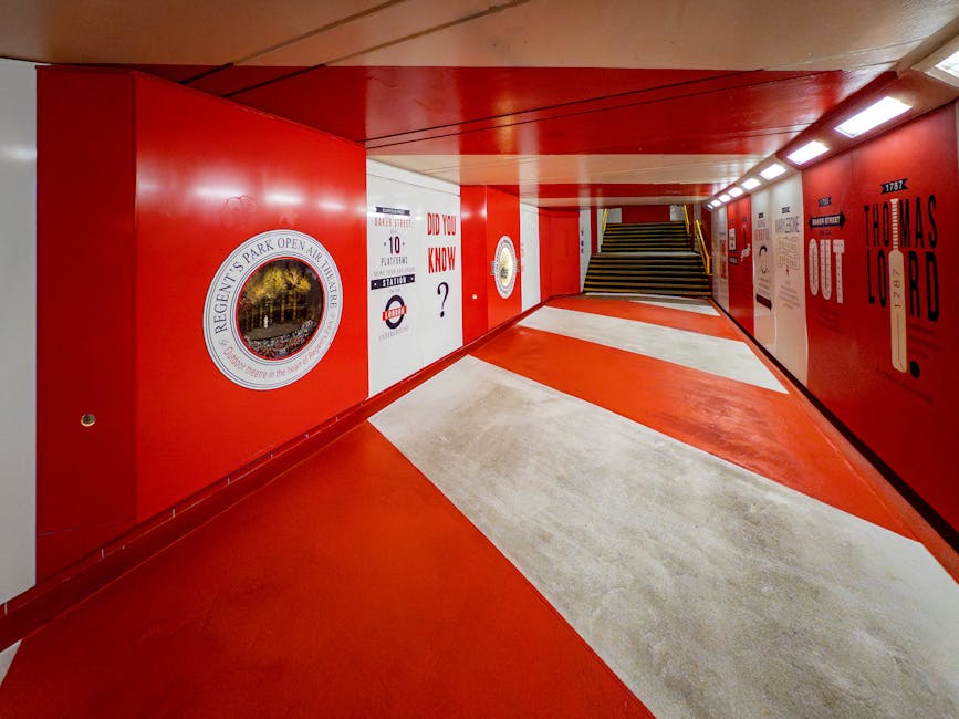 The image shows an underground passageway with a vibrant red ceiling and walls, complemented by white diagonal markings on the floor. The right wall features various posters and advertisements, includ