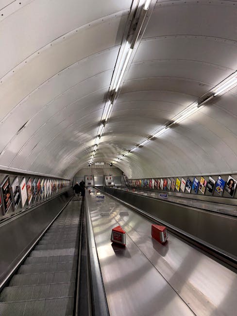 The image depicts an underground passageway with a curved white ceiling illuminated by evenly spaced fluorescent lights. Along the sides, there are colorful advertising posters, and at the far end, es