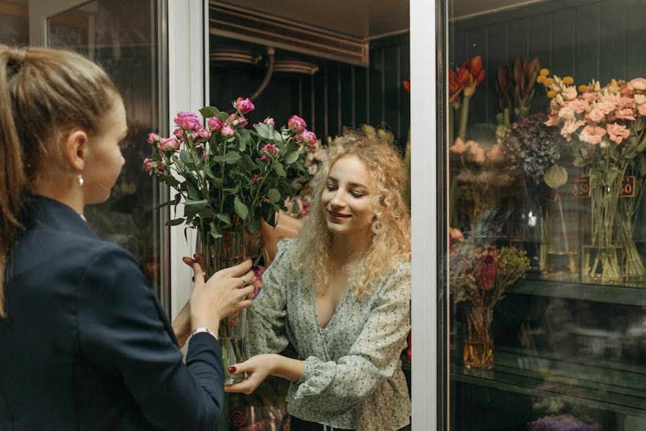 A young woman with curly blonde hair, dressed in a light-colored patterned blouse, is smiling as she receives a bouquet of fresh pink roses from a florist dressed in a dark blazer. The bouquet, held w