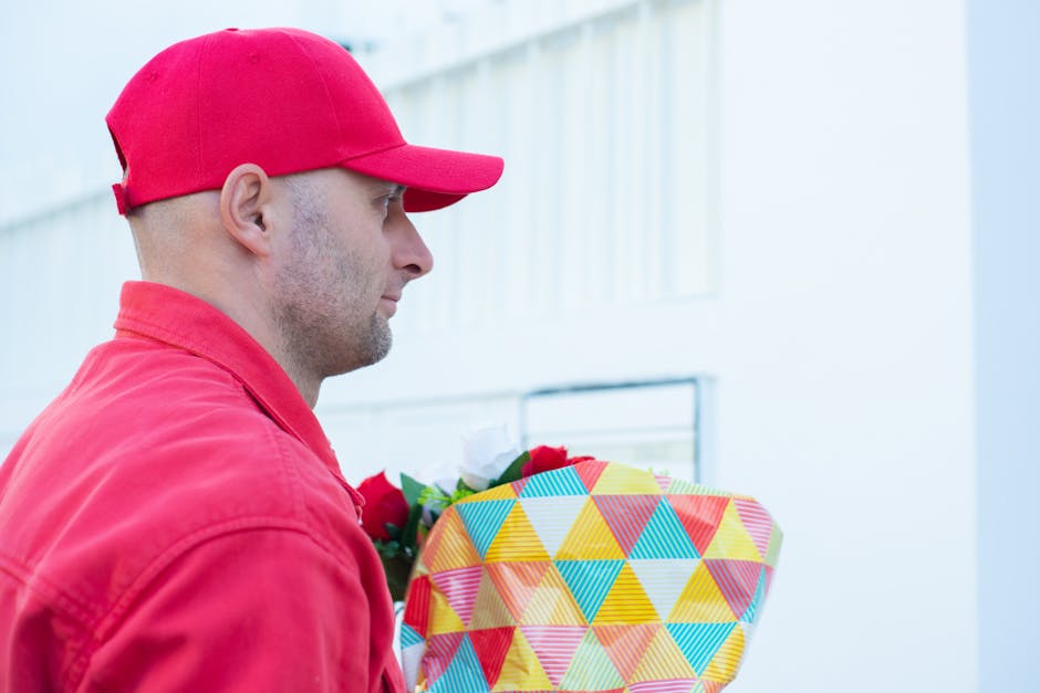 A delivery person dressed in a red cap and red jacket is holding a bouquet of fresh flowers, wrapped in colorful geometric patterned paper, outside a building with a light-colored exterior. The bouque