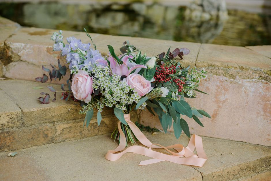 A delicate floral bouquet placed on a stone ledge outdoors, featuring soft pink roses, light purple delphiniums, white astilbes, and sprigs of red berries, complemented by eucalyptus and other green f